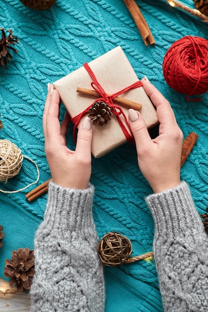 Female hands holding Christmas gift box against red socks, a blue knitted sweater, and a garland with cones.