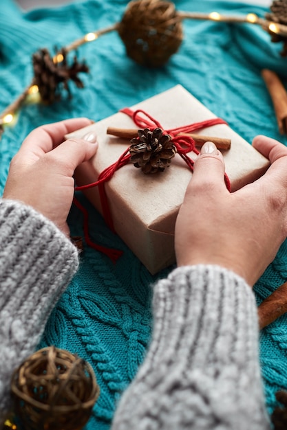 Female hands holding Christmas gift box against red socks, a blue knitted sweater, and a garland with cones.