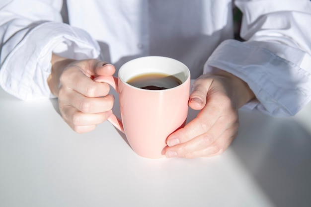 Female hands holding a cup of coffee on a white table under the morning sun