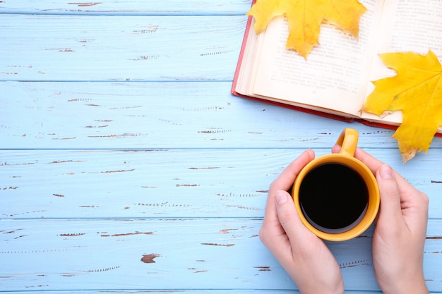 秋の紅葉と青いテーブルの上の本とブラックコーヒーのカップを保持している女性の手