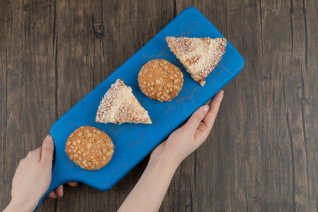 Female hands holding cutting board of wholegrain cookies and sweet pie on wooden table. 