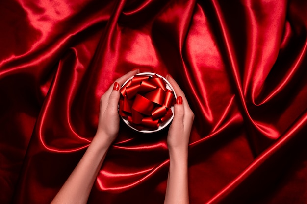 Female hands holding a festive box with ribbons and bows. Red festive background top horizontal view copyspace.