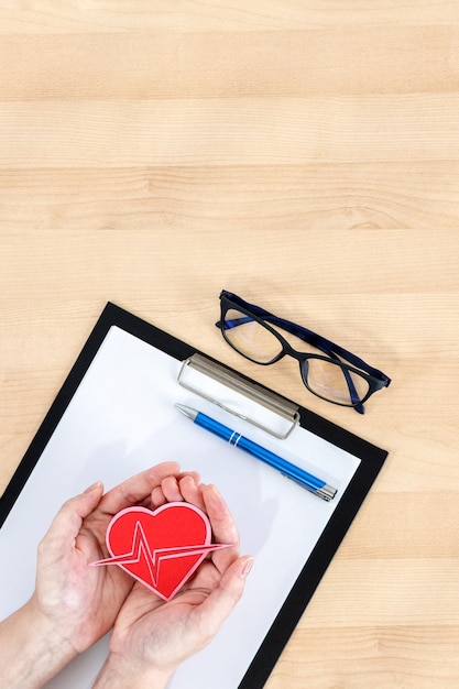 Female hands holding heart clipboard and glasses on wooden desk Top view