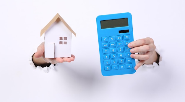 Female hands holding house figure and calculator through torn hole white paper