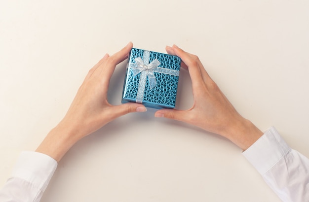 Female hands holding little blue gift box on white table, top view