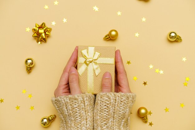 Женские руки, держащие рождественскую подарочную коробку на желтом украшенном праздничном фоне, женщина готовит ...