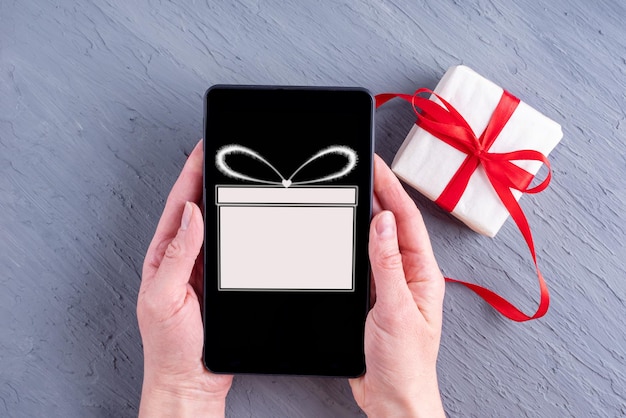 Female hands holding a phone with a gift icon and a purchased gift on a gray background. Online store application on the screen of a digital smartphone. Online sale. Online shopping concept