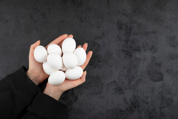 Female hands holding pile of raw eggs on black background.