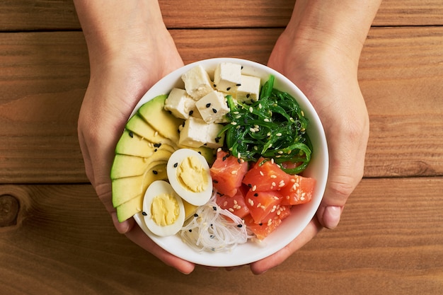 Female hands holding poke bowl with salmon, avocado on wooden background top view