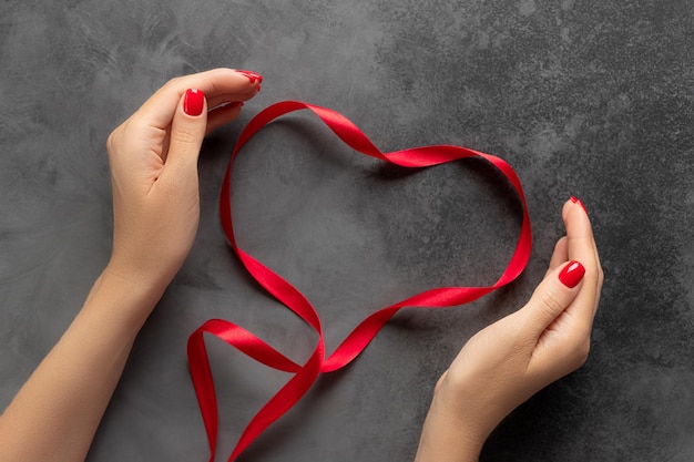 Foto mani femminili che tengono il cuore del nastro rosso su sfondo scuro