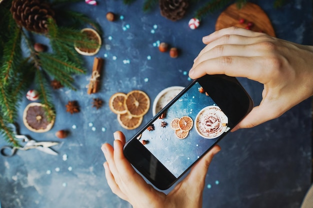 Foto mani femminili che tengono smartphone su sfondo di pietra scura con decorazione natalizia tazza di abete di cacao e arance secche natale e capodanno composizione stile di vita blogger vista dall'alto messa a fuoco selettiva