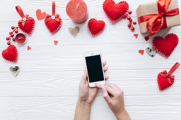 Mani femminili che tengono smartphone su un tavolo bianco con cuori decorativi per san valentino.