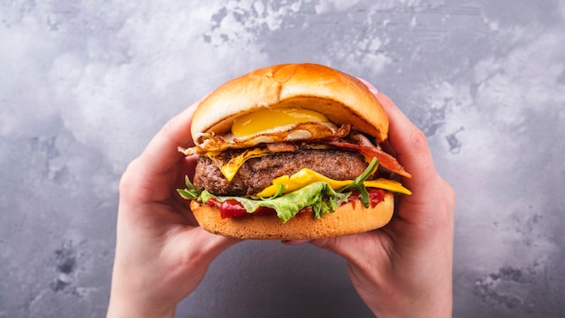 Mani femminili che tengono gustosi hamburger di manzo con uova fritte e pancetta vista dall'alto cibo americano