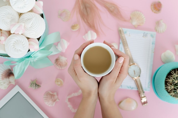 Mani femminili che tengono una tazza bianca con bevanda sul fondo di rosa pastello.