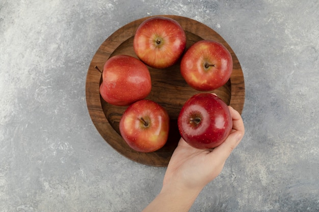 Female hands holding wooden plate of red apples on marble surface. 
