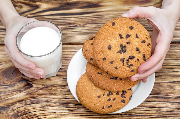 Female hands holds glass of milk and chocolate chip cookies over wooden table