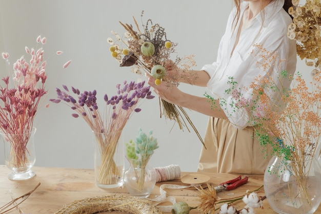 Mani femminili che fanno un bouquet eterno di fiori secchi su un tavolo di legno con un bouquet di fiori e decorazioni nel suo studio del negozio di fiori