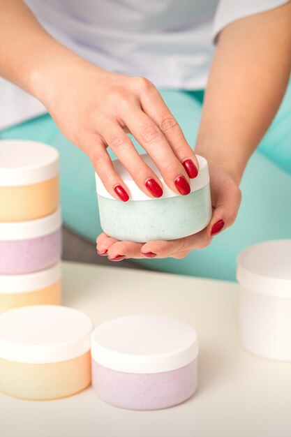 Female hands open a white body cream jar over the table with many jars of cosmetics.