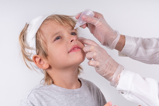 Female hands optometrist wearing transparent medical gloves putting eye drop patient little blonde boy eyes in ophthalmology clinic. Sick look. White background.