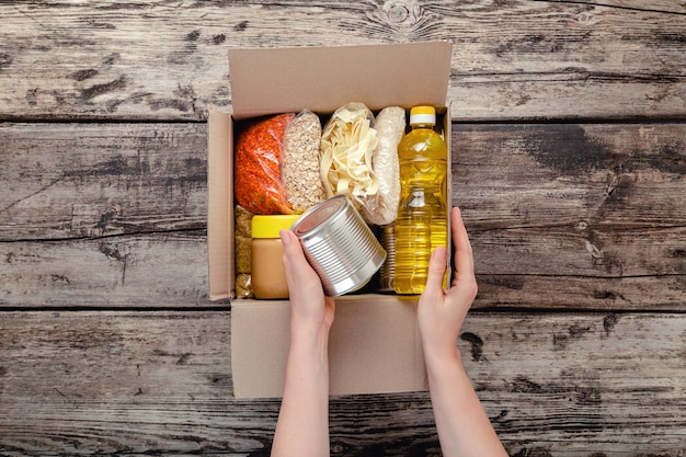 Female hands packing donation box with food items of staple products on wooden table. Person woman receiving donation Food box. Donate Food delivery concept. Donations grocery canned food. Top view.