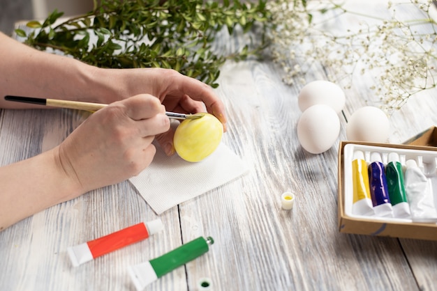 Female hands painting an egg in yellow with a brush on a wooden table. Preparing for Easter