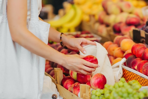 女性の手は、食品市場で果物や野菜を綿花の農産物バッグに入れます。ショッピング用の再利用可能なエコバッグ。ゼロウェイストのコンセプト。