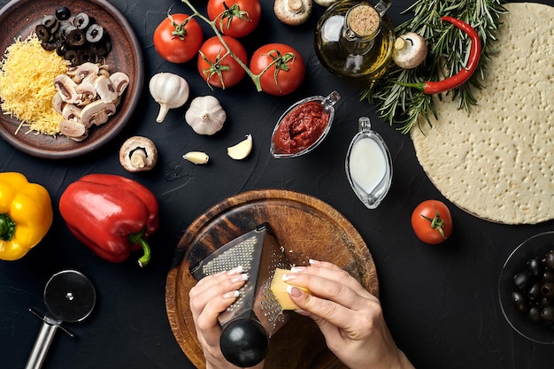 Female hands rubbed cheese grated on pizza ingredients for cooking pizza on black table top view