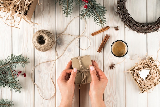 Female hands tie bow on gift box in festive package top view Spruce branches cinnamon cup of coffee and twine ball Crafting present Objects layout Christmas or New Year holiday concept