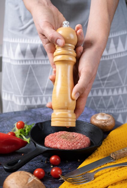 Female hands use a wooden mill for spices pray pepper on a meat\
patty raw in a cast iron pan