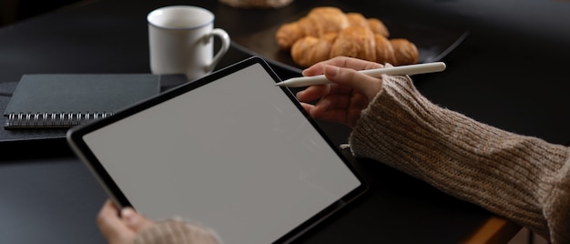 Female hands using tablet with stylus pen on breakfast table with schedule books, coffee and croissant