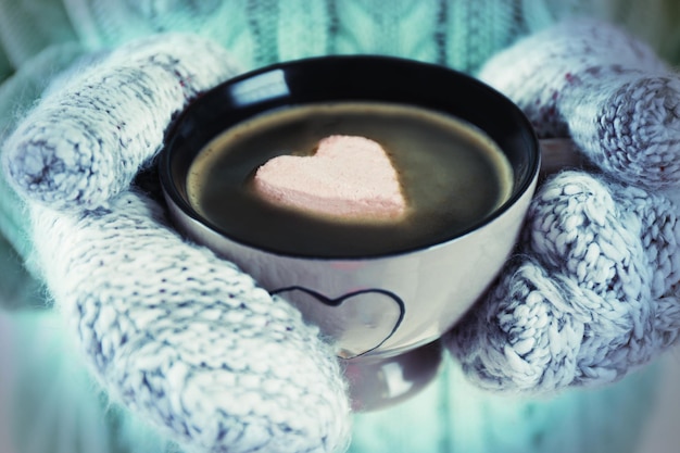 Female hands in warm mittens holding cup of hot coffee with heart marshmallow close up