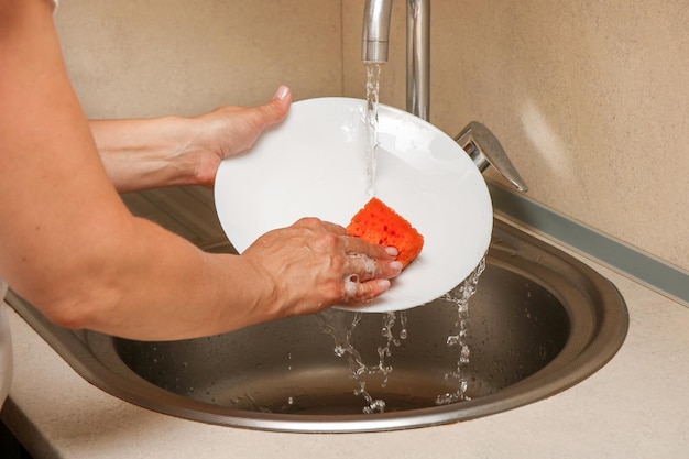 female hands wash a plate with a dish sponge under running tap water