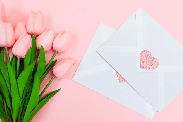 Female hands with a bouquet of pink tulips and blank white letter envelopes, on a pink background. View from above.