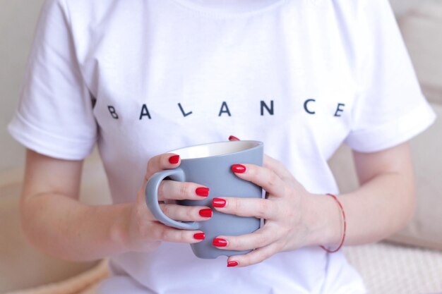 빨간 매니큐어가 있는 여성의 손은 비문 균형이 있는 흰색 티셔츠를 들고 있습니다.