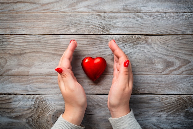 Female hands with red nails and decorative heart on wooden background.