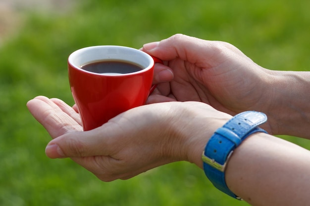 手首の時計を持つ女性の手は、背景に緑の草とコーヒーの赤い磁器のカップを保持します。カップに選択的な焦点。