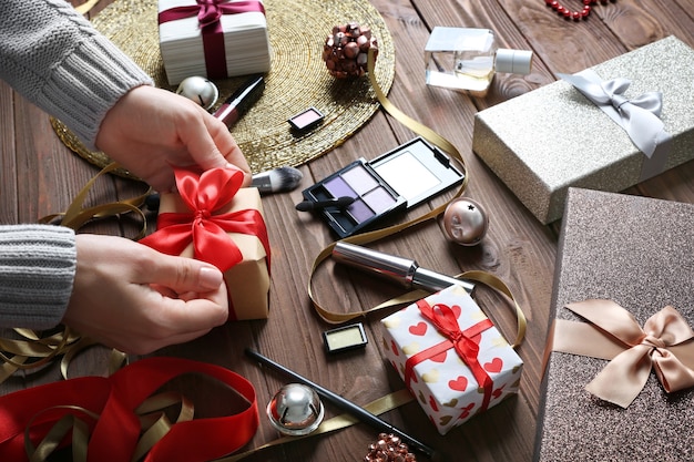 Female hands wrapping Christmas gift and colorful makeup cosmetic on wooden surface