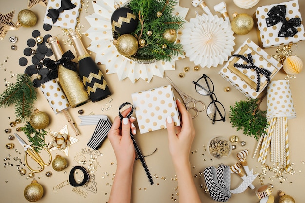 Female hands wrapping gifts on desk with Christmas decoration background in golden and black colors. Flat lay, top view