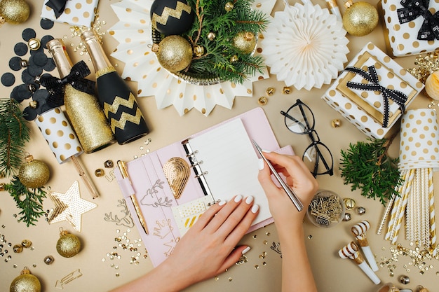 Женские руки пишут в ежедневнике или блокноте. Новогоднее украшение фон в золотых и черных тонах. Плоская планировка, вид сверху