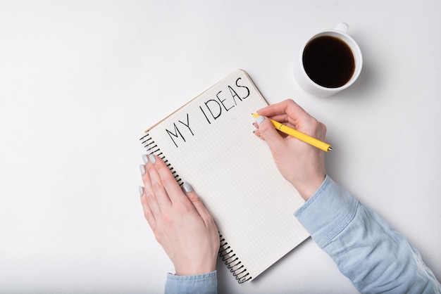 Female hands writing words MY IDEAS Top view of Notepad and cup of coffee white background