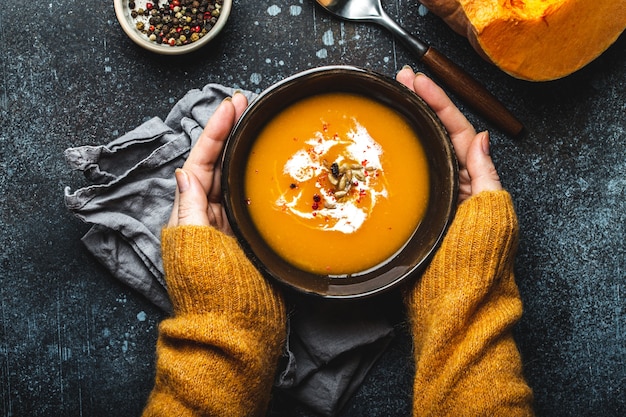 Photo female hands in yellow knitted sweater holding a bowl with pumpkin cream soup on dark stone background with spoon decorated with cut fresh pumpkin, top view. autumn cozy dinner concept
