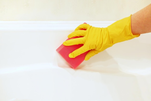 A female handyellow rubber glove with a pink sponge washes polishes the surface white bathtub