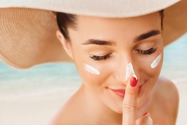 Female in hat applying moisturizing lotion on skin Beautiful Young woman with sun cream on face