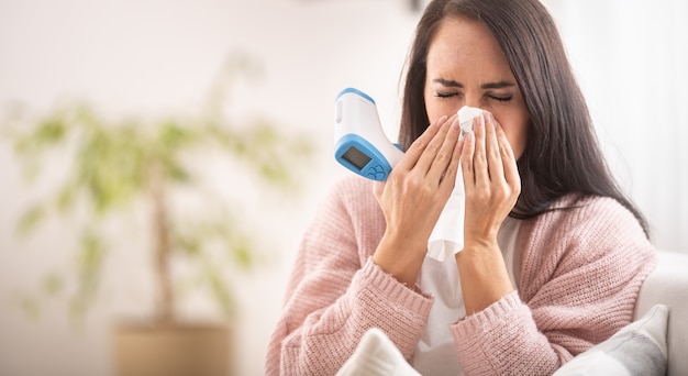 Female having flu or cold sneezes into a tissue and holds a digital thermometer.