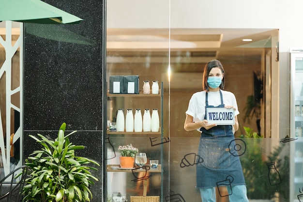 Female healthy food store owner in medical mask showing welcome sign