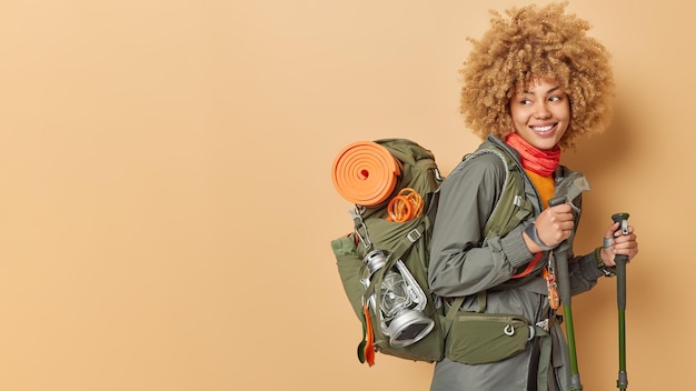 Female hiker poses with backpack trekking poles wears sports wear looks happily away isolated over beige background mock up space for your advertising content Active tourist reaches top of mountain