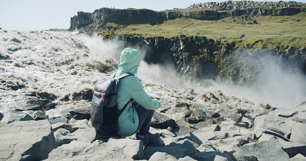 Photo female hiker with backpack sitting at cliff edge enjoying detifoss waterfall in iceland