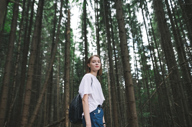 Female hiker with a backpack standing in the mountain coniferous forest looking at camera