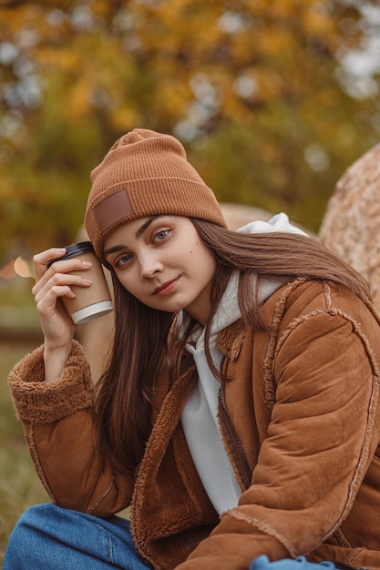 Female hipster with cup of takeaway coffee sitting near rocks in nature and chilling in autumn park