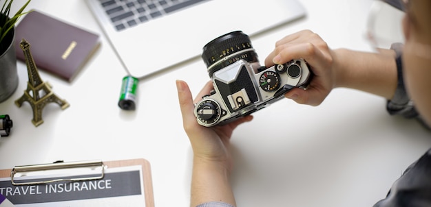 Female holding camera while preparing travel items on white table with laptop and travel insurance form